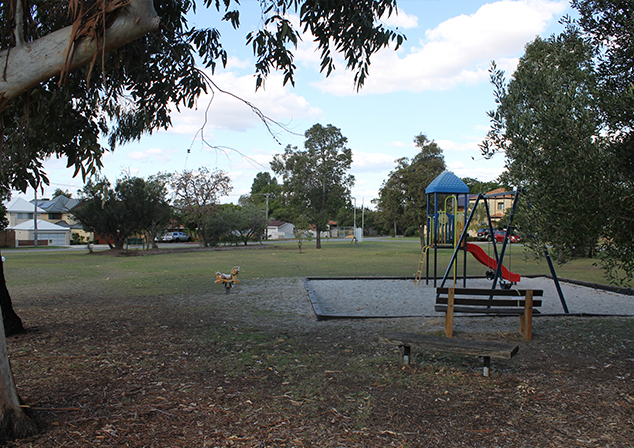 Photo of dirt area that transitions into grass. Sandpit with playground equipment and park benches in area between grass and dirt. Trees in background.