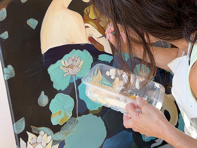 A woman stands with her back to the camera as she paints a light cream coloured acrylic onto a canvas with turquoise flowers.