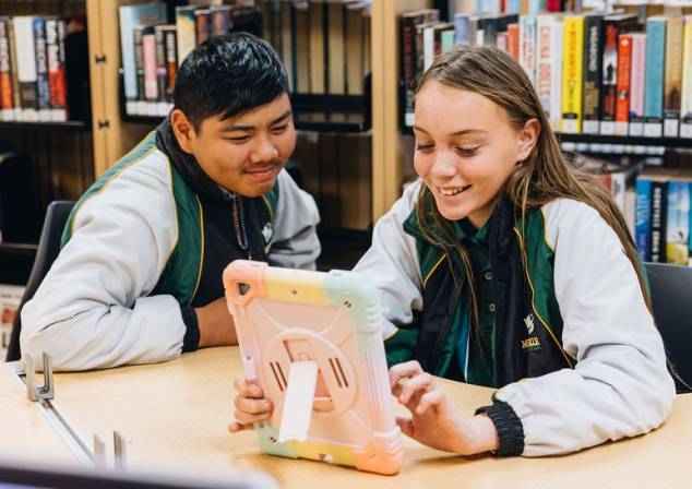 Two students sit together at a table, viewing an iPad and smiling.