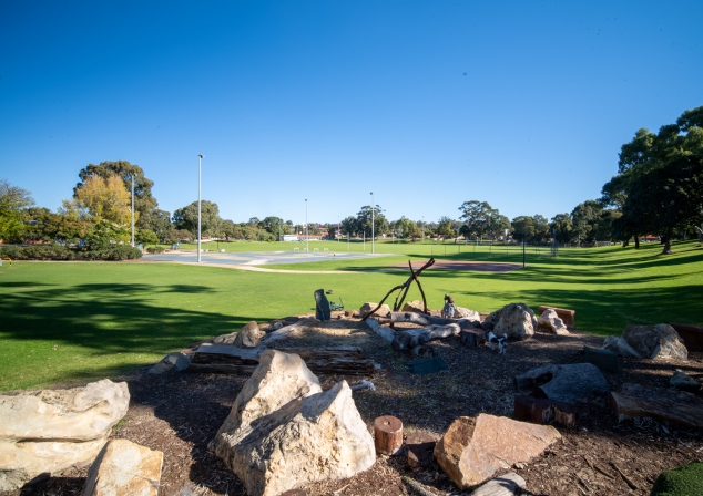 Photo of grassy area with two concrete slabs and trees in background, and mulch pit with rocks and boulders in it in foreground.